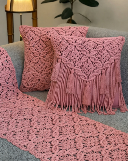 A close-up view of a dusty rose handcrafted macrame home decor set on a grey sofa, showcasing a fully knotted table/sofa runner, one all-over patterned cushion cover, and one fringe-draped cushion cover decorated with hanging tassels