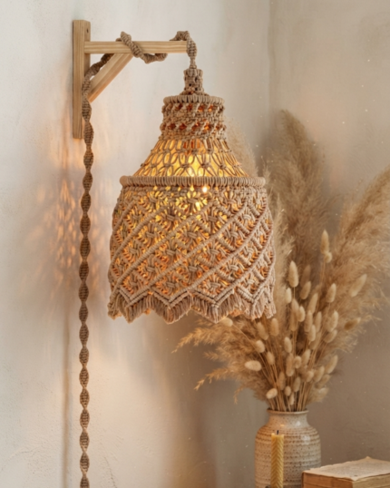"A cozy room corner displaying a lit handcrafted boho macrame wall hanging pendant light suspended from an L-shaped wooden wall bracket, with a completely braided cotton cord cover and a textured wave-patterned shade, next to a vase with dried pampas grass."