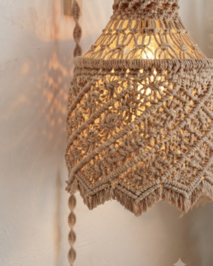 "A cozy room corner displaying a lit handcrafted boho macrame wall hanging pendant light suspended from an L-shaped wooden wall bracket, with a completely braided cotton cord cover and a textured wave-patterned shade, next to a vase with dried pampas grass."