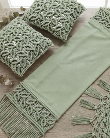 An overhead flat lay of an organic sage green macrame home decor set on a wooden table, showcasing a fabric table runner with leaf-patterned macrame borders, two matching textured cushion covers, and a circular fringed coaster next to a cup