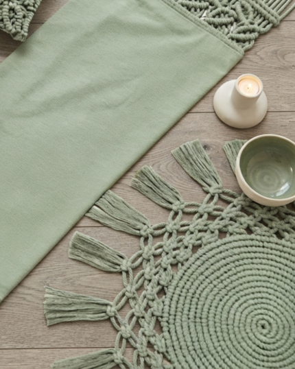 An overhead flat lay of an organic sage green macrame home decor set on a wooden table, showcasing a fabric table runner with leaf-patterned macrame borders, two matching textured cushion covers, and a circular fringed coaster next to a cup