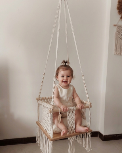 A frontal view of a handcrafted boho-chic macrame baby swing hanging indoors, featuring a secure hand-knotted cream cotton basket, rounded wood security bars, a plush off-white cushion, and flowing cotton tassels below the seat."