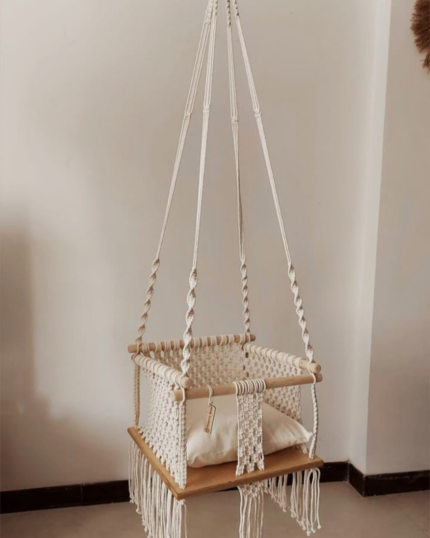 A frontal view of a handcrafted boho-chic macrame baby swing hanging indoors, featuring a secure hand-knotted cream cotton basket, rounded wood security bars, a plush off-white cushion, and flowing cotton tassels below the seat."