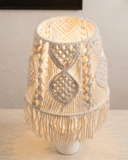 A side-angle view of an lit handcrafted bohemian macrame table lamp featuring a hand-knotted cream cotton shade with wooden bead trim, a fringe border, and a white cylindrical ceramic base, set on a stone countertop."