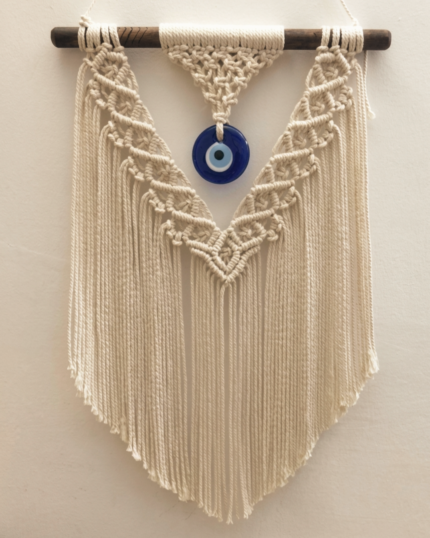 A frontal view of a handcrafted cream macrame evil eye wall hanging suspended from a dark wooden dowel on a beige wall, featuring a central blue glass Nazar charm and detailed geometric V-shaped fringes."