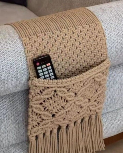 A close-up view of a handcrafted beige macrame sofa armrest organizer draped over a grey textured couch arm, featuring a detailed hand-knotted pocket holding a black TV remote control, finished with elegant tassels hanging below."