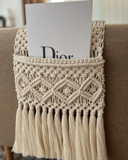 A close-up view of a handcrafted beige macrame sofa armrest organizer draped over a grey textured couch arm, featuring a detailed hand-knotted pocket holding a black TV remote control, finished with elegant tassels hanging below."