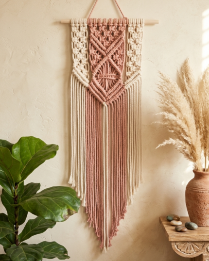 A frontal view of a handcrafted dual-tone blush pink and cream macrame wall hanging on a smooth wooden dowel, displayed on a textured beige wall next to a potted green plant and dried pampas grass.